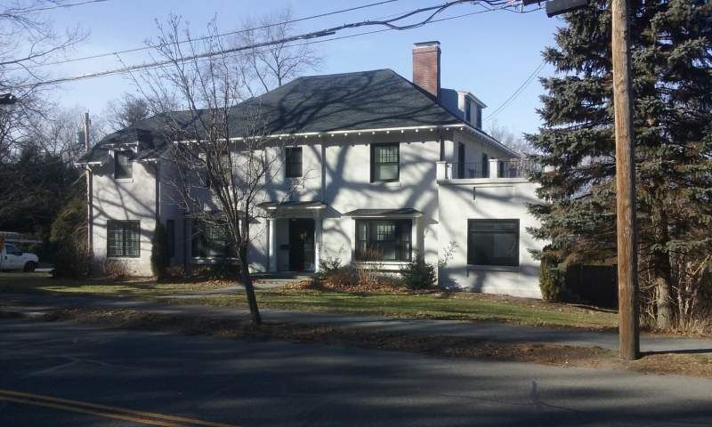 Two-story white house with black roof, chimney, and dark windows, viewed from across a street.