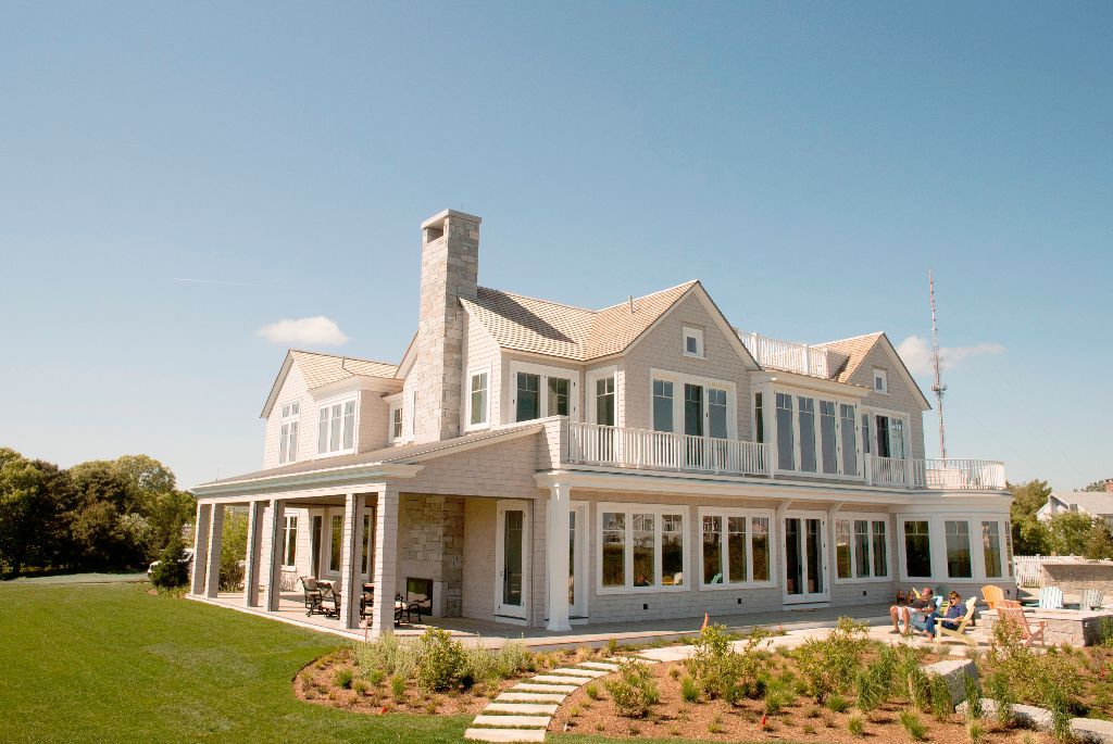 Large, multi-story house with a stone facade and a wraparound porch, set on a green lawn with a clear blue sky.
