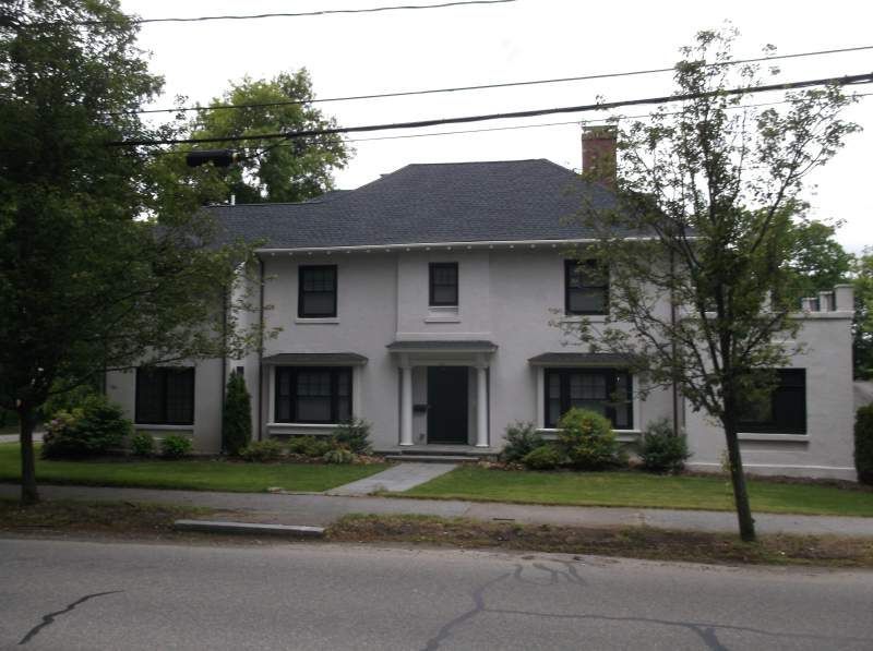Two-story stucco house with black-framed windows, dark roof, and a front walkway. Trees and sky visible.