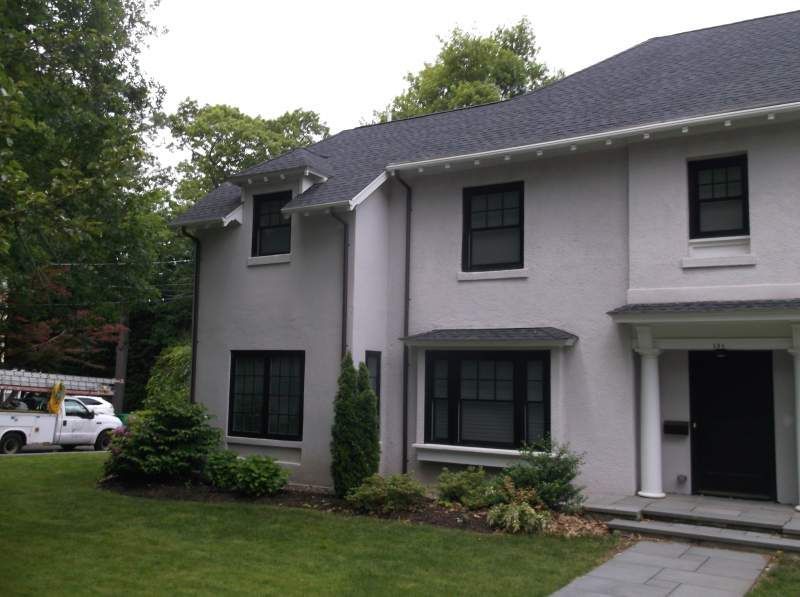 Two-story house with light-colored stucco exterior, black windows, dark roof, and green lawn.