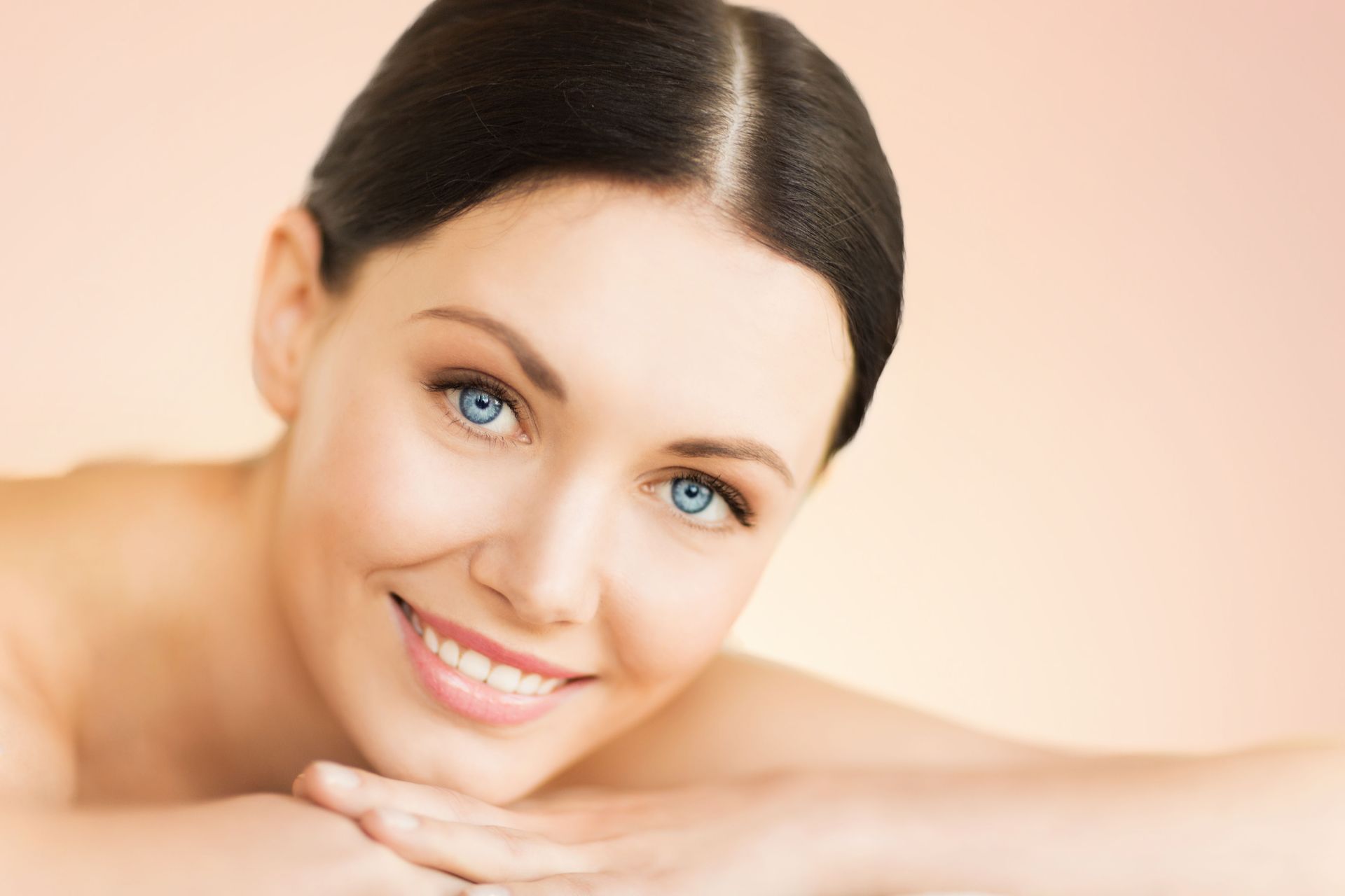 Woman with blue eyes smiling, resting arms, soft lighting, peach background.
