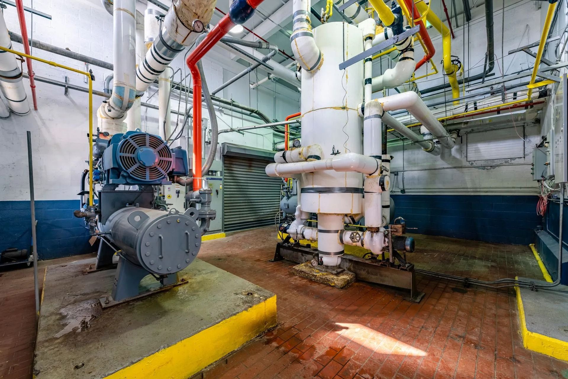 Industrial mechanical room with pipes, tanks, and machinery; red, yellow, and blue piping; dirty floor.