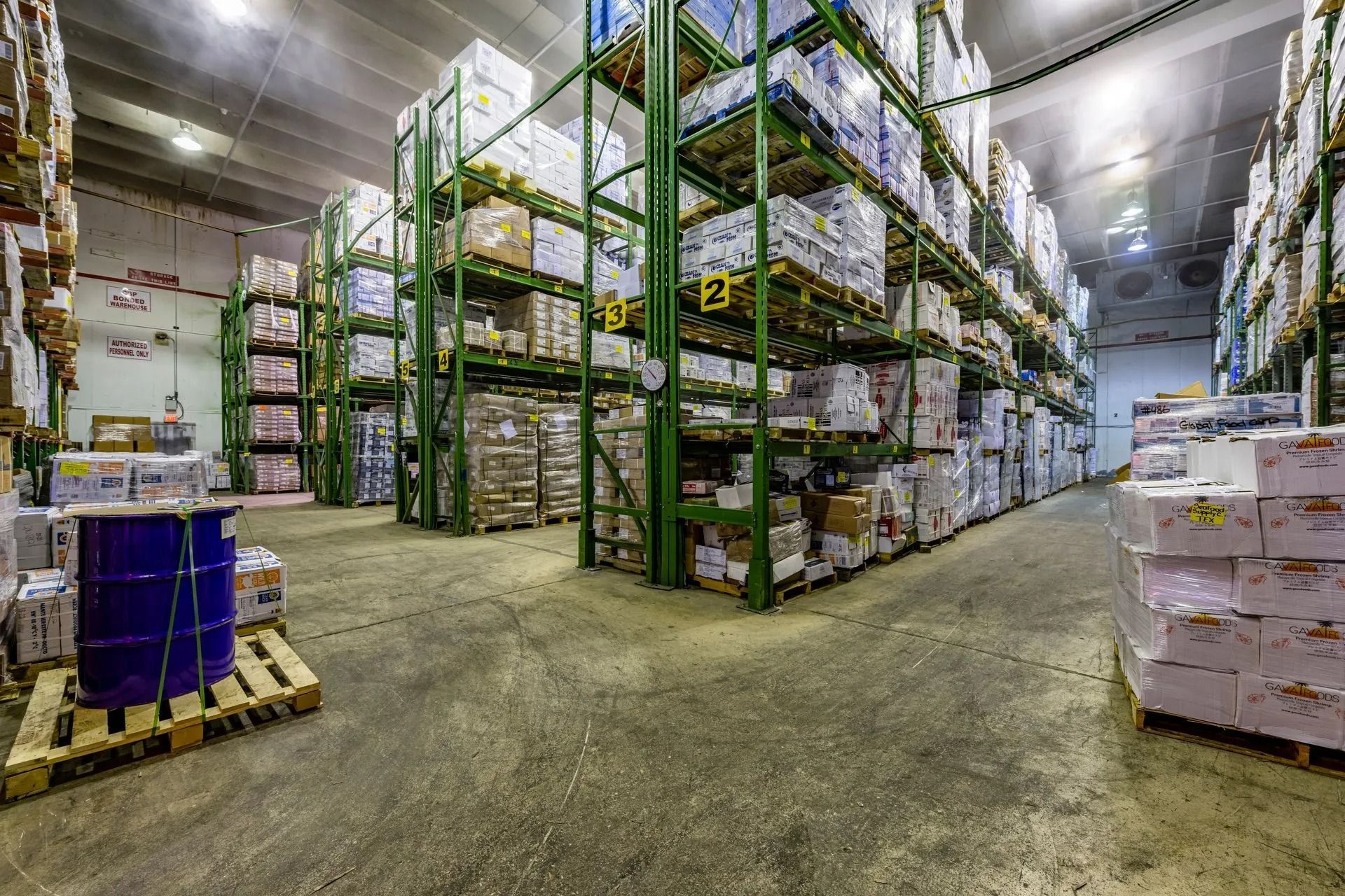 Inside a cold storage warehouse: stacked pallets with boxed goods, fluorescent lighting, and metal shelving.