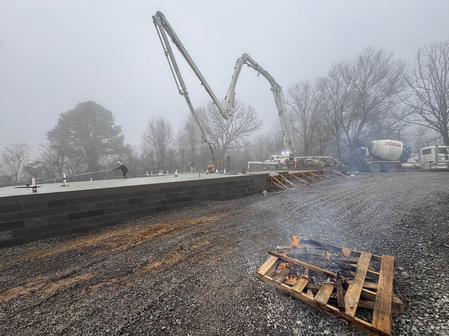 Construction site with a concrete pump pouring concrete on a slab in a foggy outdoor setting.