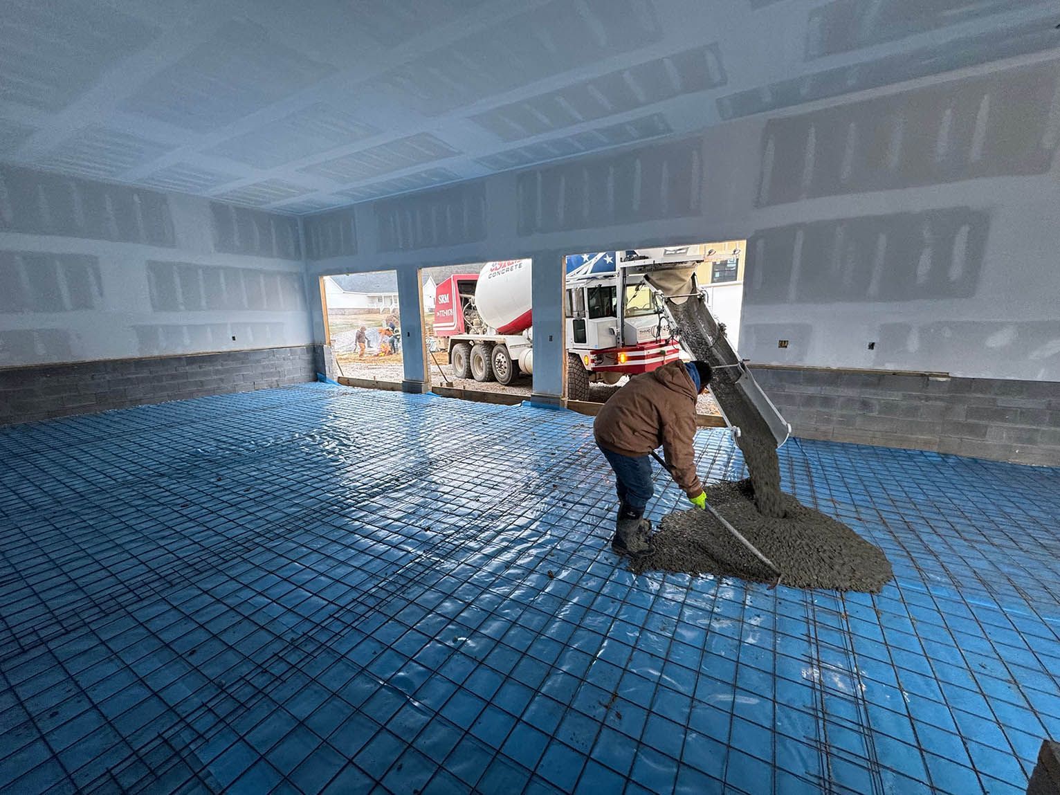 Concrete being poured on a blue membrane over rebar inside a building, construction worker.
