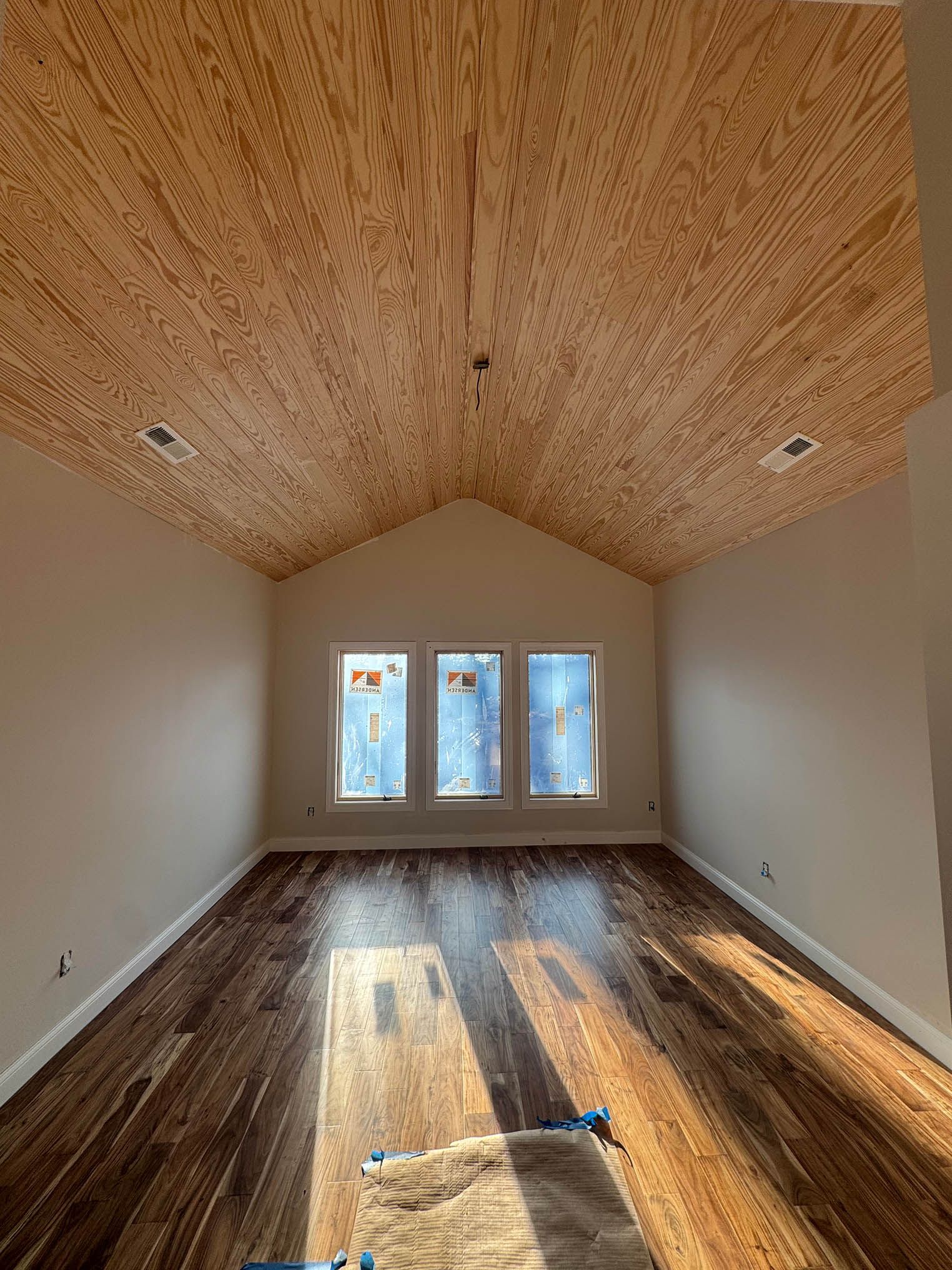 Room with wood-paneled ceiling, three windows, and wood-look flooring. Light streams in; walls are white.