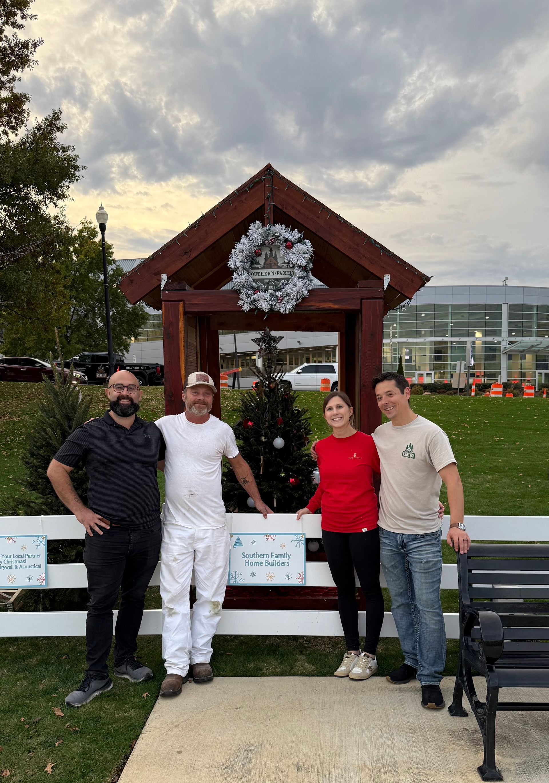 Four people pose by a small wooden structure with a Christmas tree and a wreath in an outdoor setting.