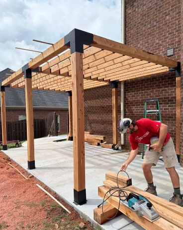 A person building a wooden pergola on a concrete patio next to a brick building.