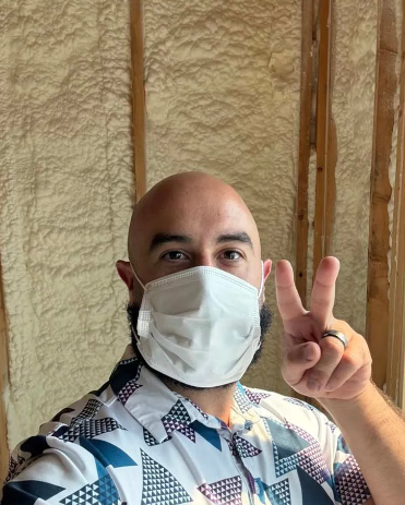 Man wearing a mask gives a peace sign, standing in a room with spray foam insulation.