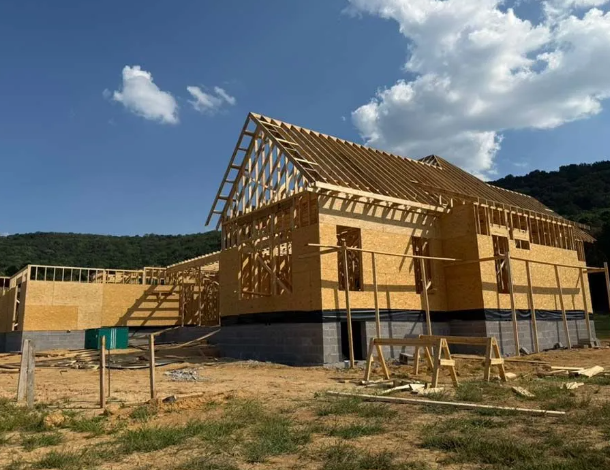 Construction of a house: wooden frame, exposed studs, partially constructed roof. Outdoors, sunny with blue sky.