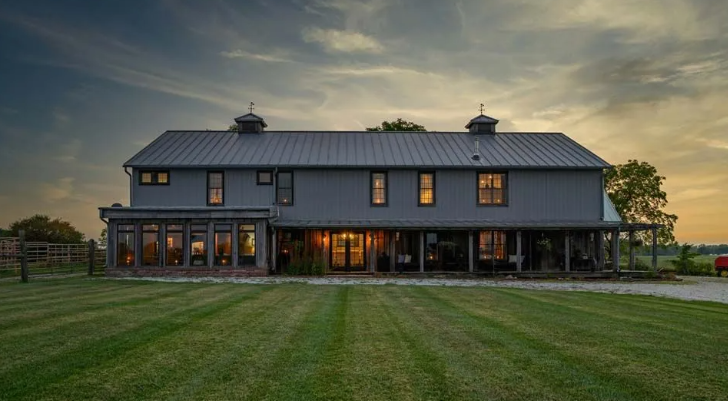 Gray farmhouse with a long front porch and green lawn under a sunset sky.