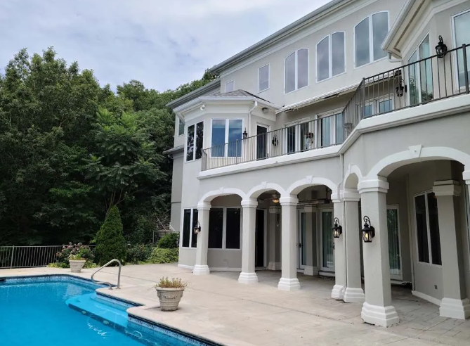 Exterior of a large, two-story house with a swimming pool. Trees are in the background, gray and white facade.