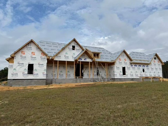 House under construction; Tyvek wrap, framing visible, partially sheathed roof.