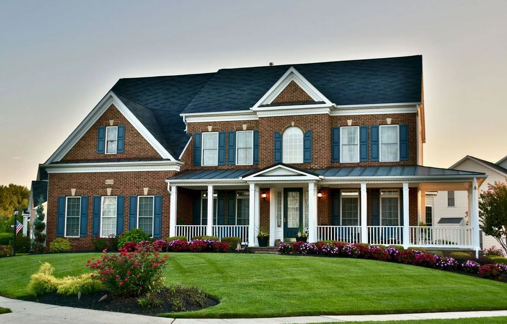 Two-story brick house with blue shutters and roof, white porch, and green lawn.