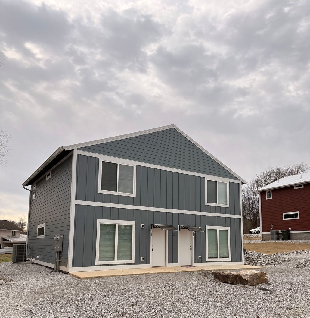 Two-story gray duplex under overcast sky; white trim, doors, and windows. Gravel yard.