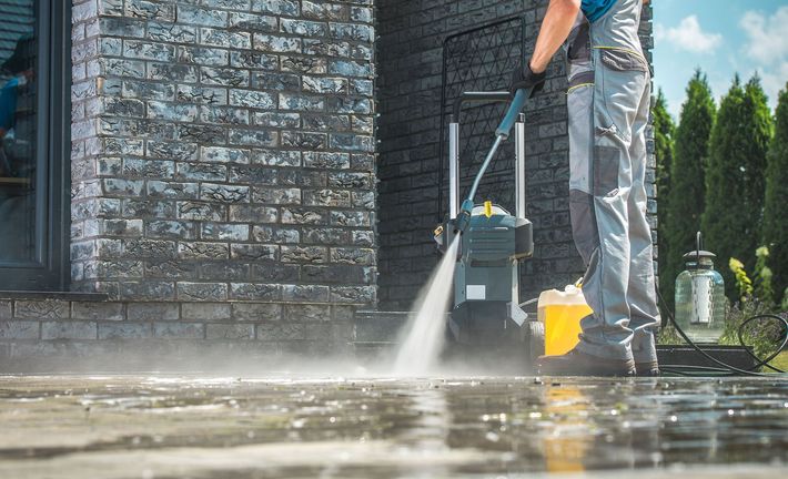 A man is using a high pressure washer to clean the sidewalk in front of a brick building
