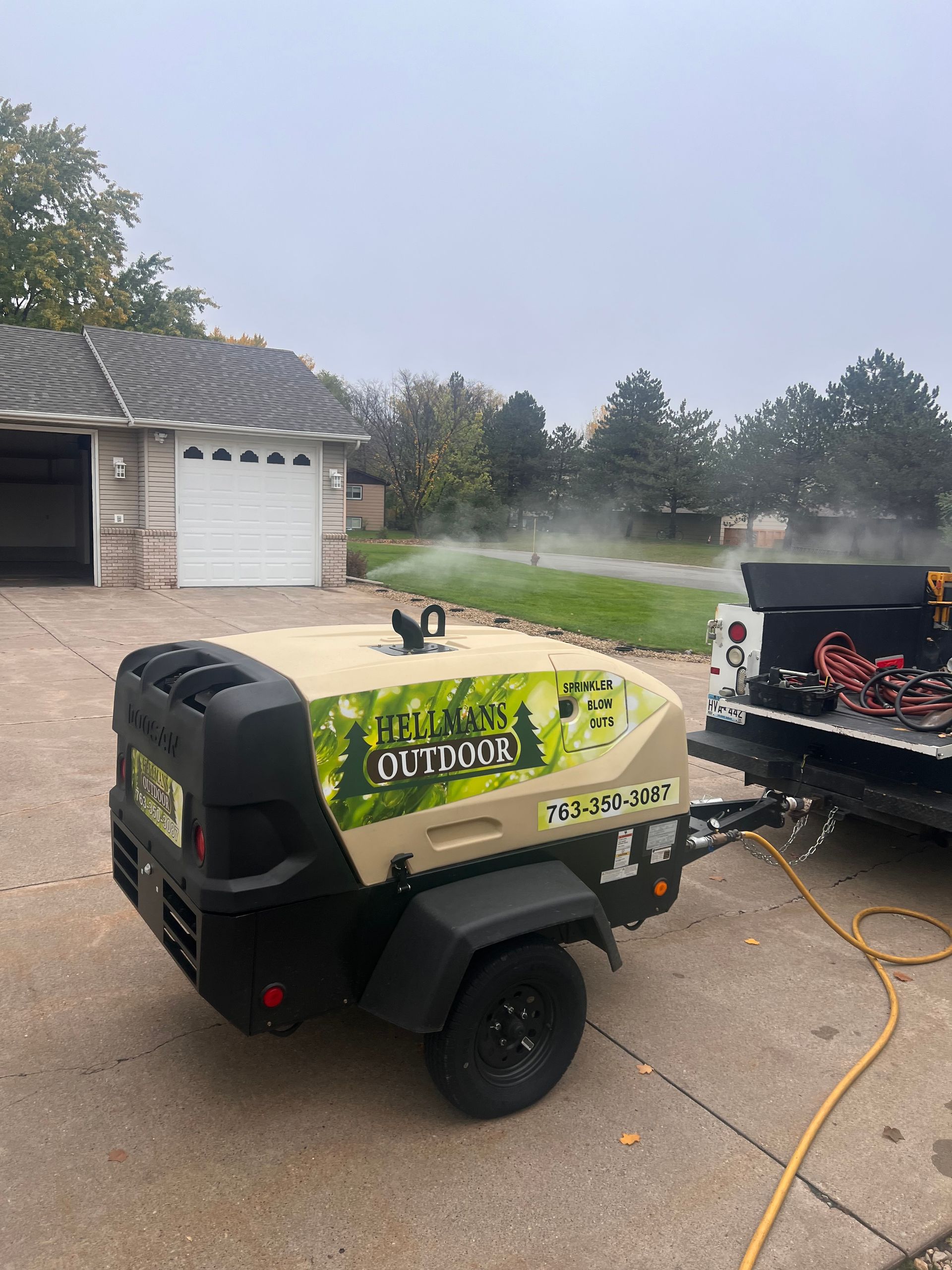 Green lawn care machine beside a black trailer spraying water in a suburban driveway