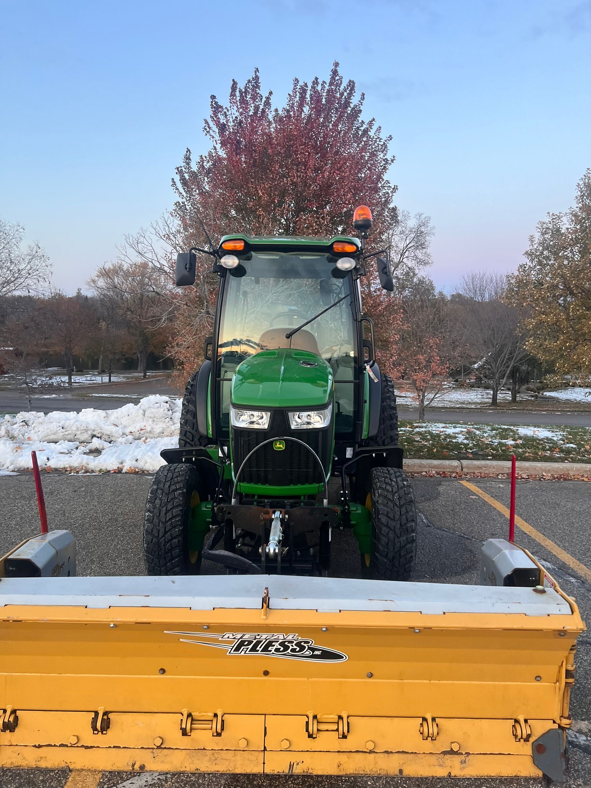 Green tractor behind a yellow snowplow in a snowy field with trees in the background