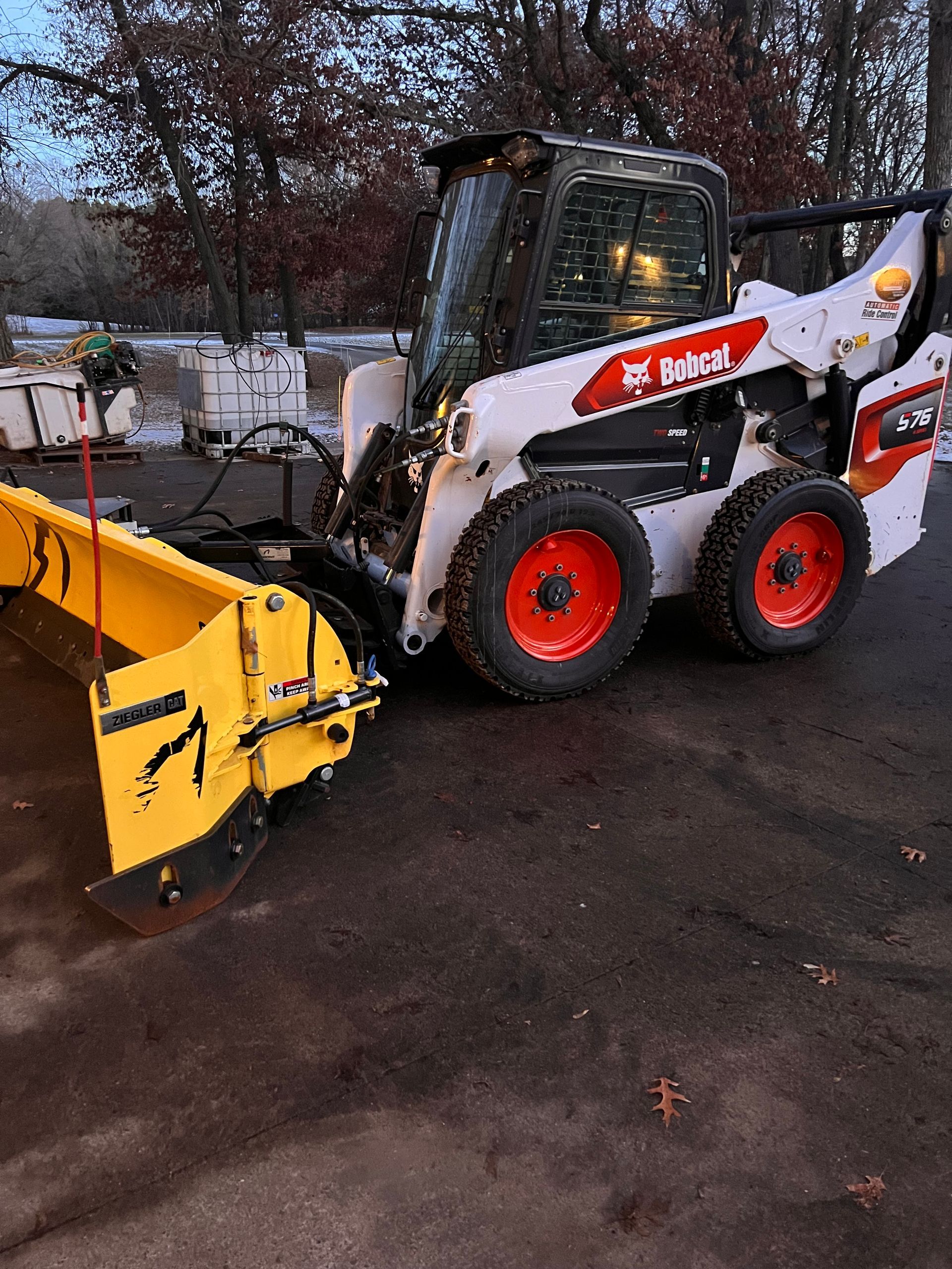 White Bobcat skid steer with red wheels and yellow snowplow attachment on wet pavement outdoors