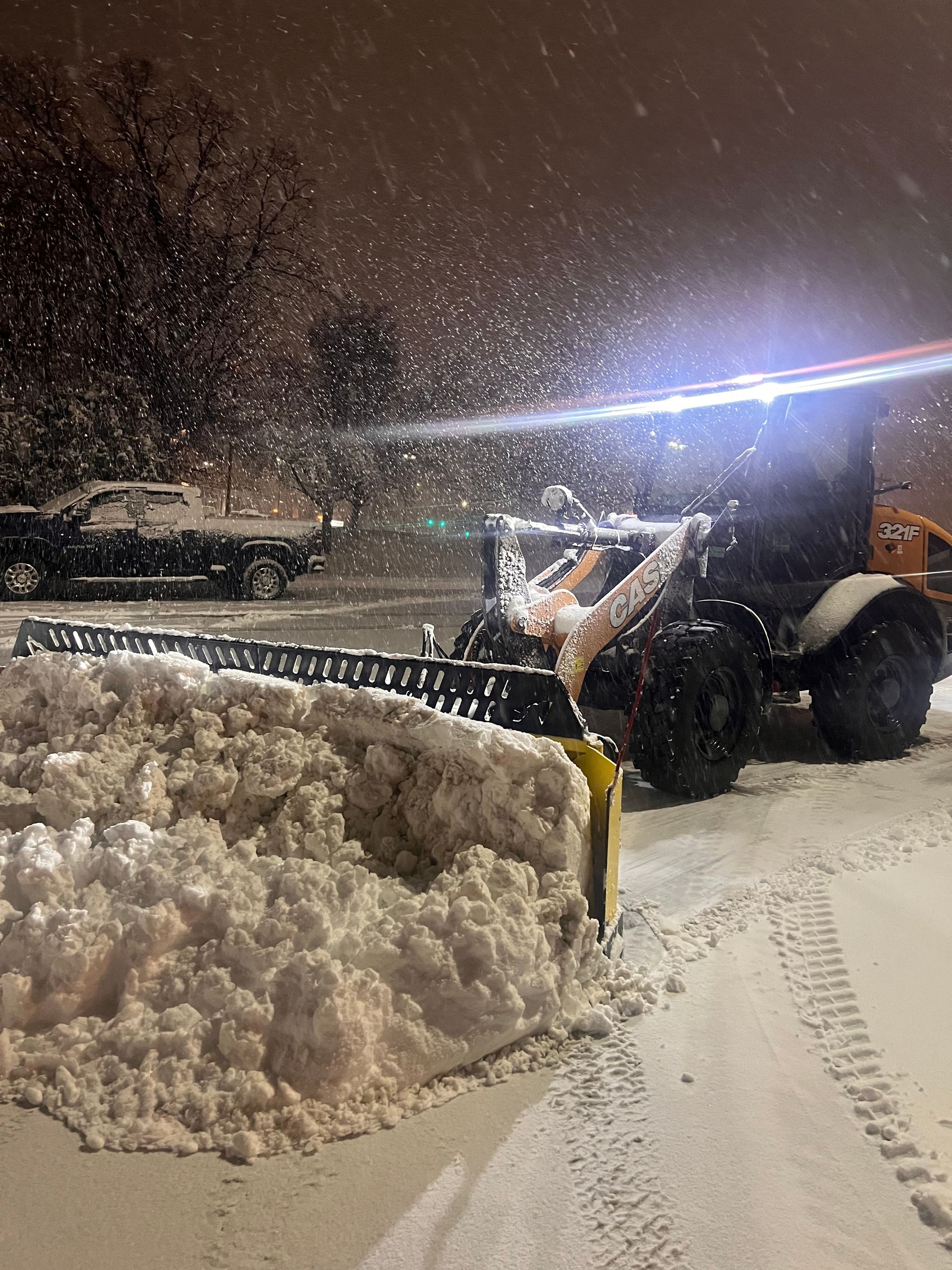 Snowplow clearing a large pile of snow at night, with bright headlights and falling snow.