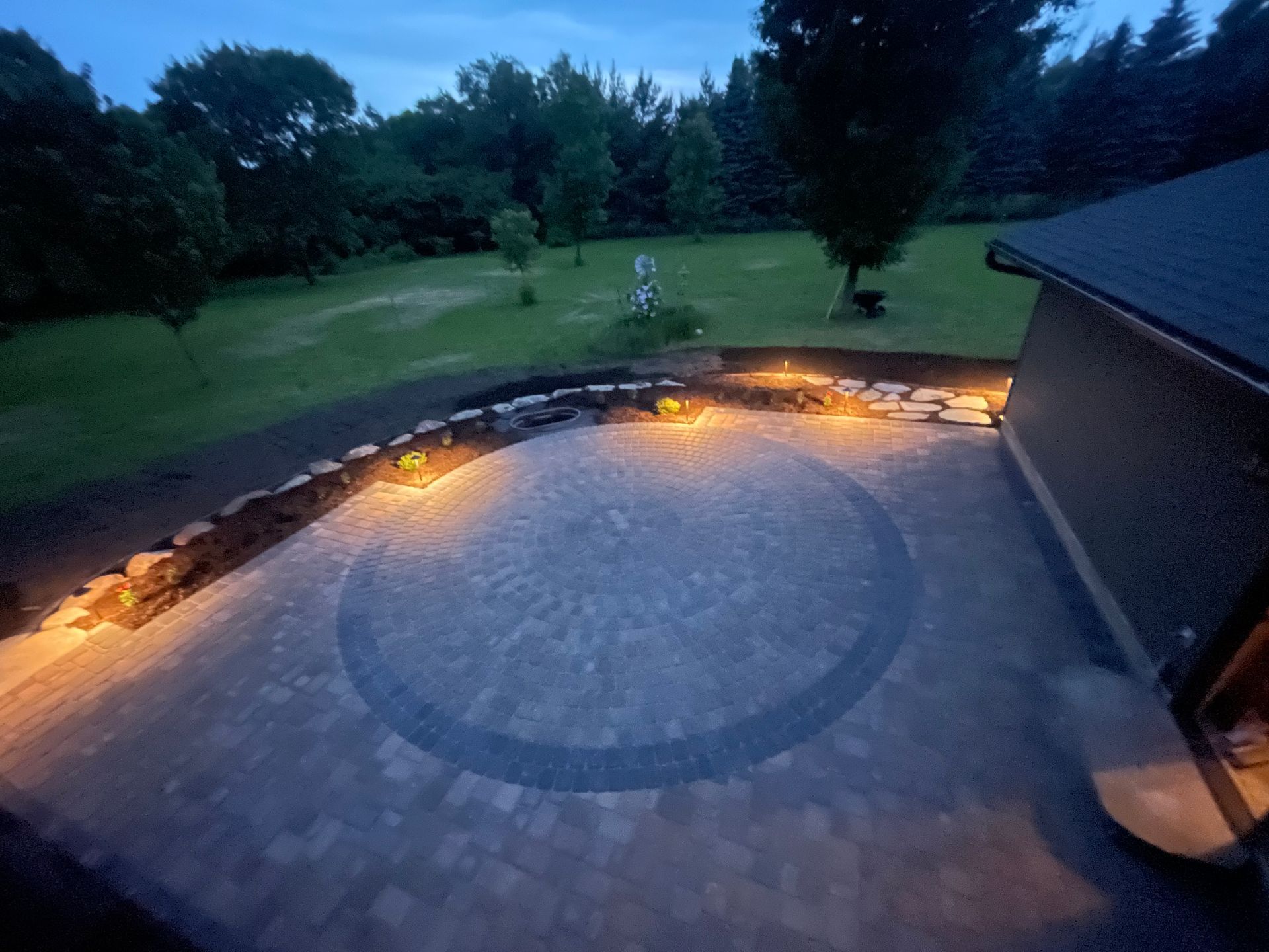 Twilight patio with stone pavers, curved fire pit seating, glowing lights, and a backyard lawn and trees.
