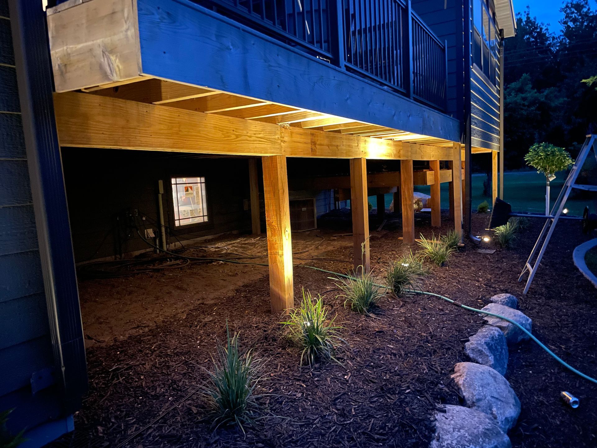 Blue building on tall wooden stilts over a dirt yard at night, with plants and large rocks nearby