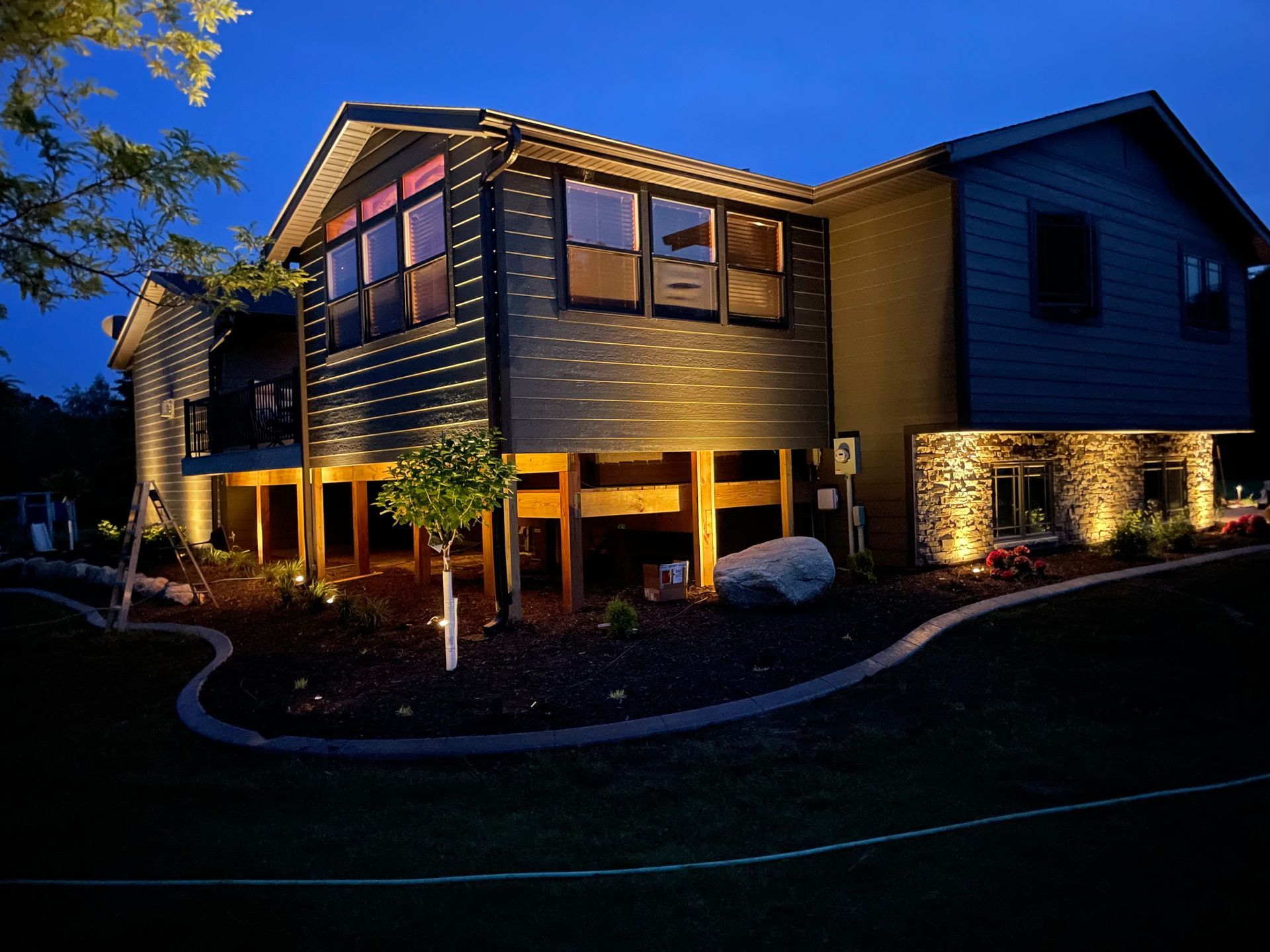 Two-story house with lit windows and exterior lights at dusk, surrounded by landscaped yard