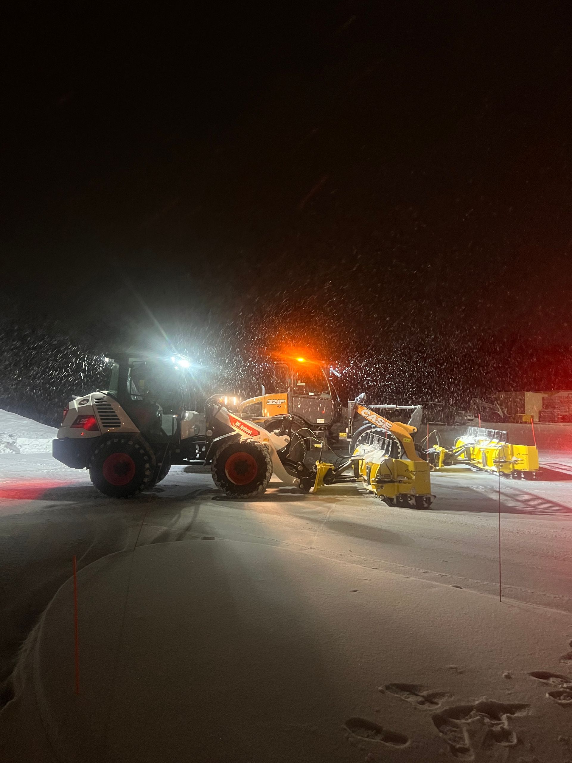 Snowplows and a loader working on a snowy road at night under bright lights