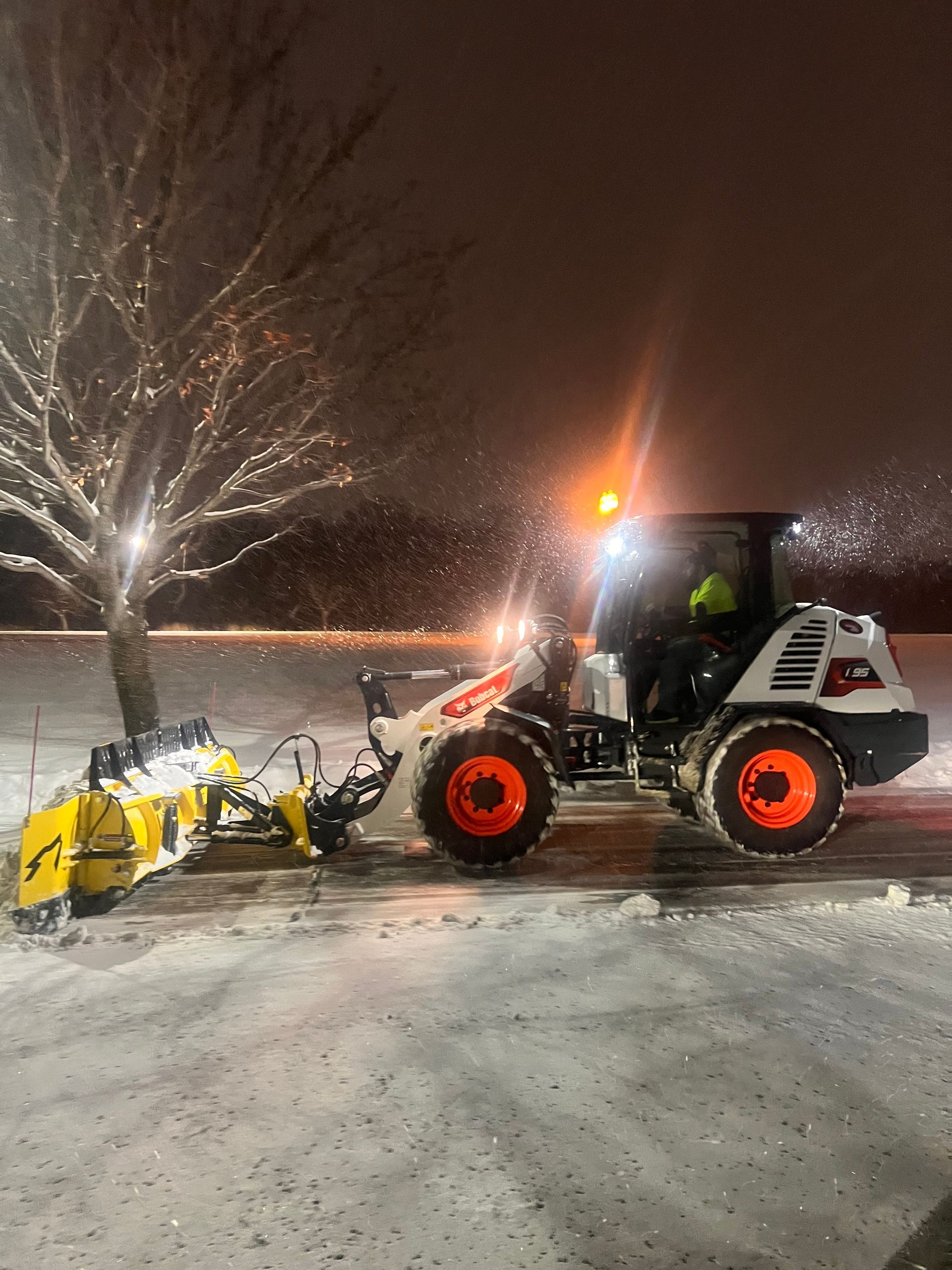 Tractor with a snowplow clearing a snowy parking lot at night under bright lights
