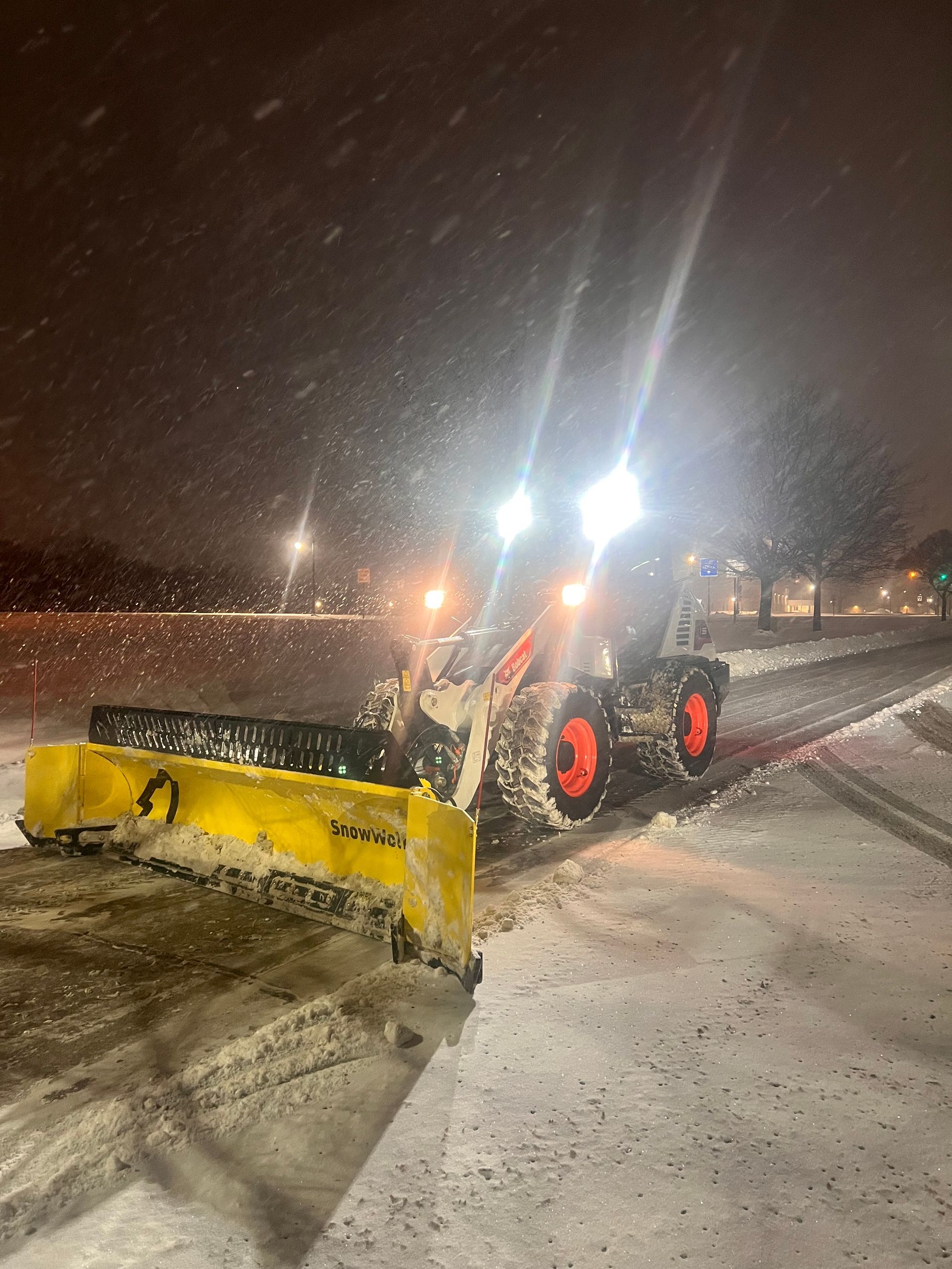 Yellow snowplow clearing a snowy road at night with bright headlights and falling snow