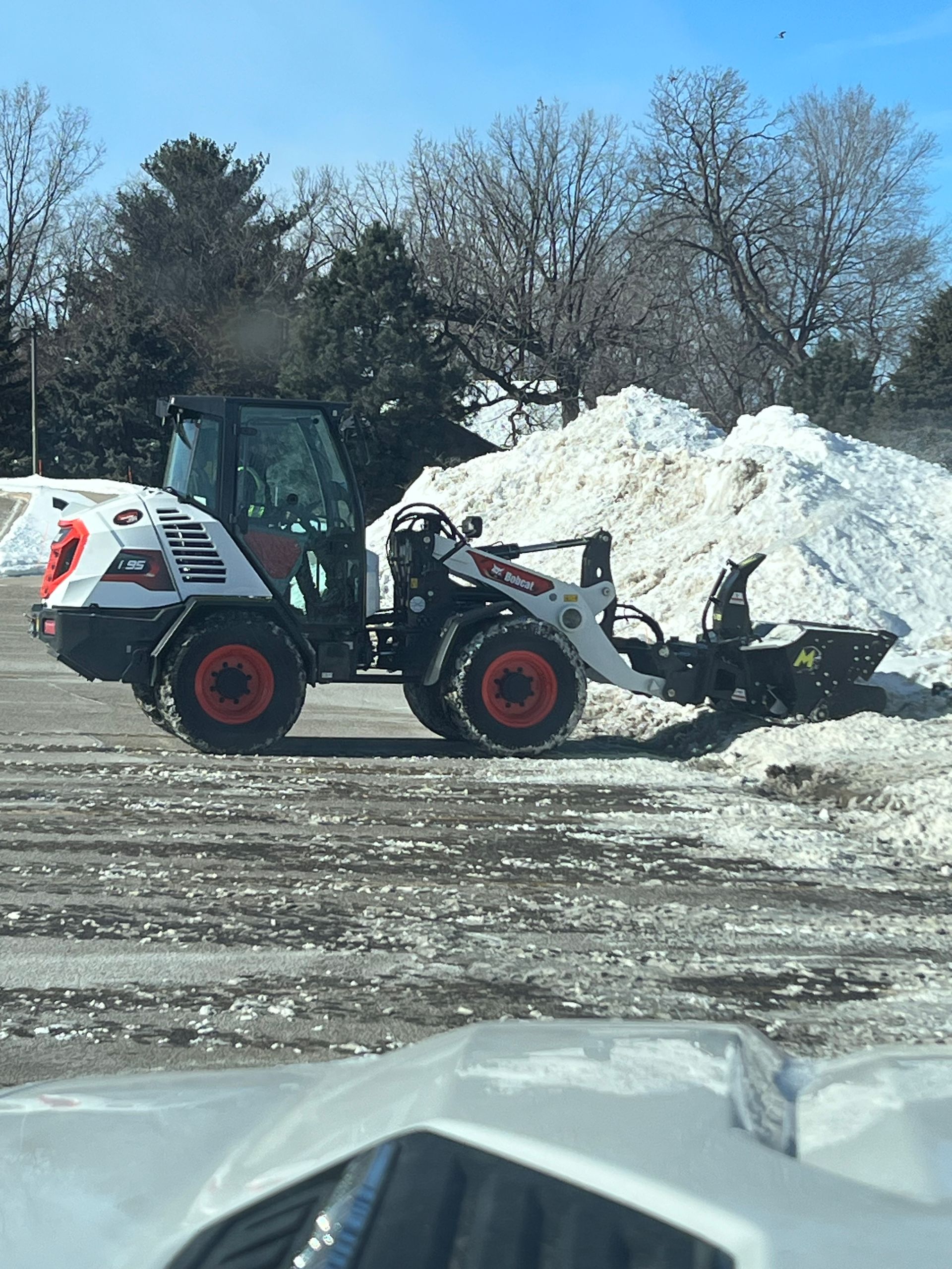 Skid steer pushing snow beside a large snow pile in a snowy lot
