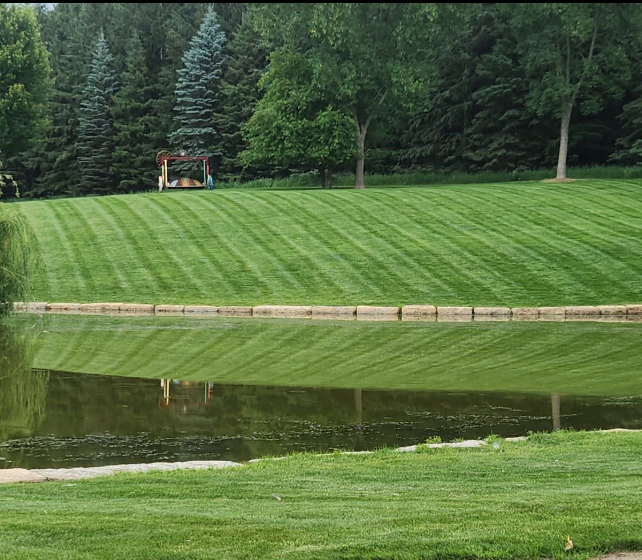 Manicured green lawn beside a calm pond, with trees and a small pavilion in the background.