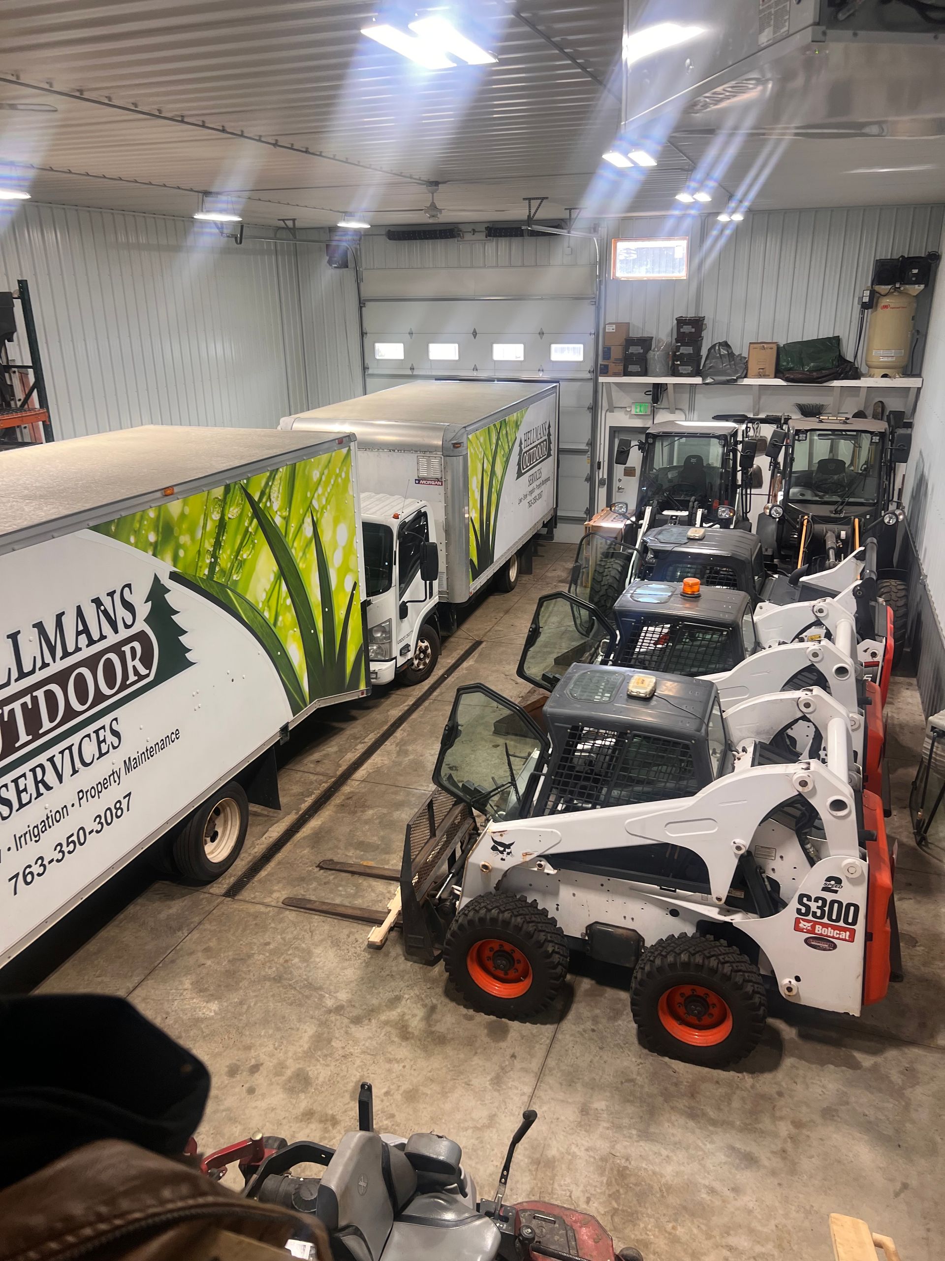 Indoor equipment showroom with several white skid steers and a large trailer featuring green graphics