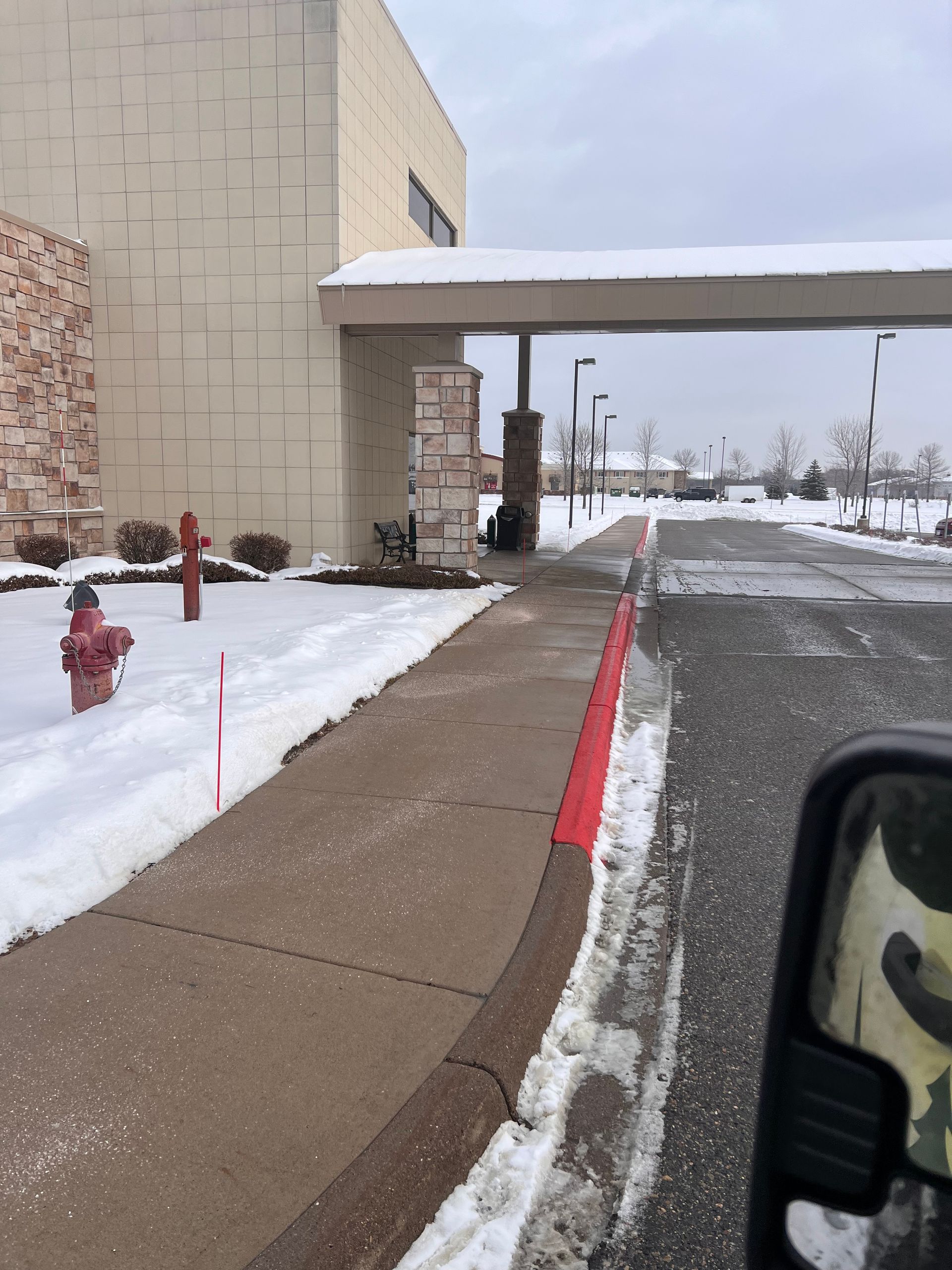 Snowy curbside sidewalk beside a beige building with a covered drive-through entrance and parked vehicle.