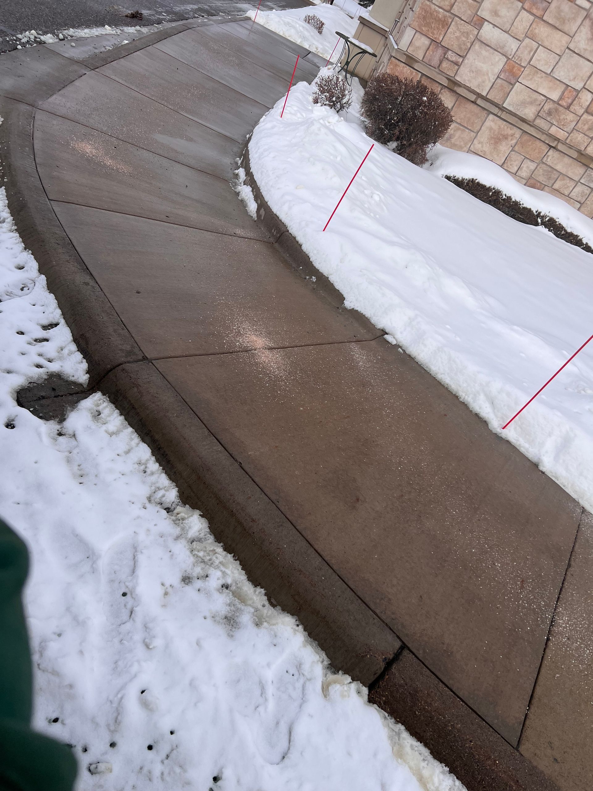 Tilted view of a snowy sidewalk beside a wet concrete path with red-topped survey markers and shrubs