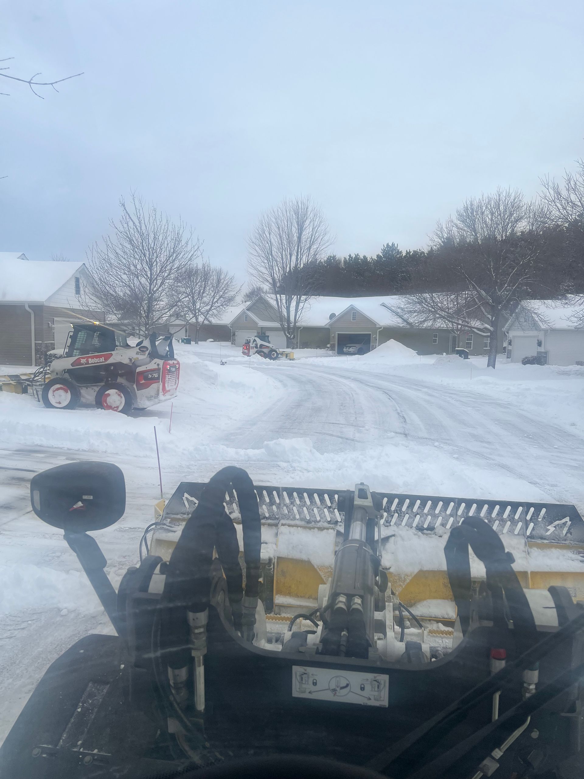 Snowplow blade clearing a snowy residential driveway with houses and trees in the background