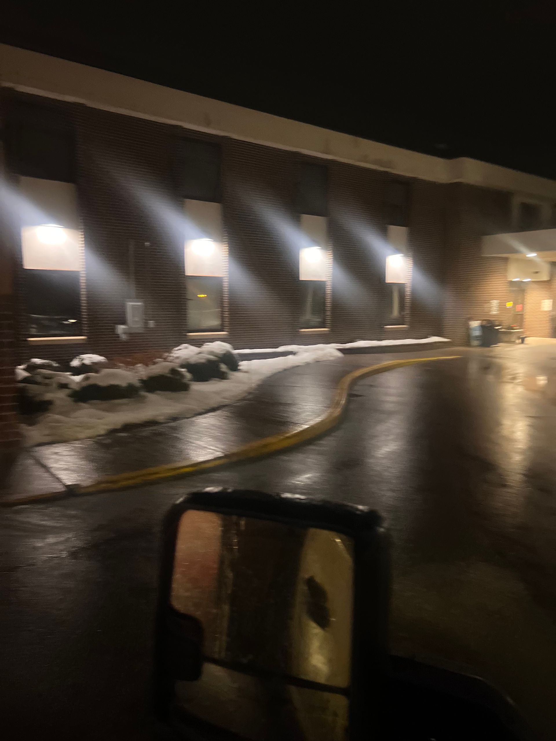 Snowy parking lot outside a lit building at night, with wet pavement reflecting lights.