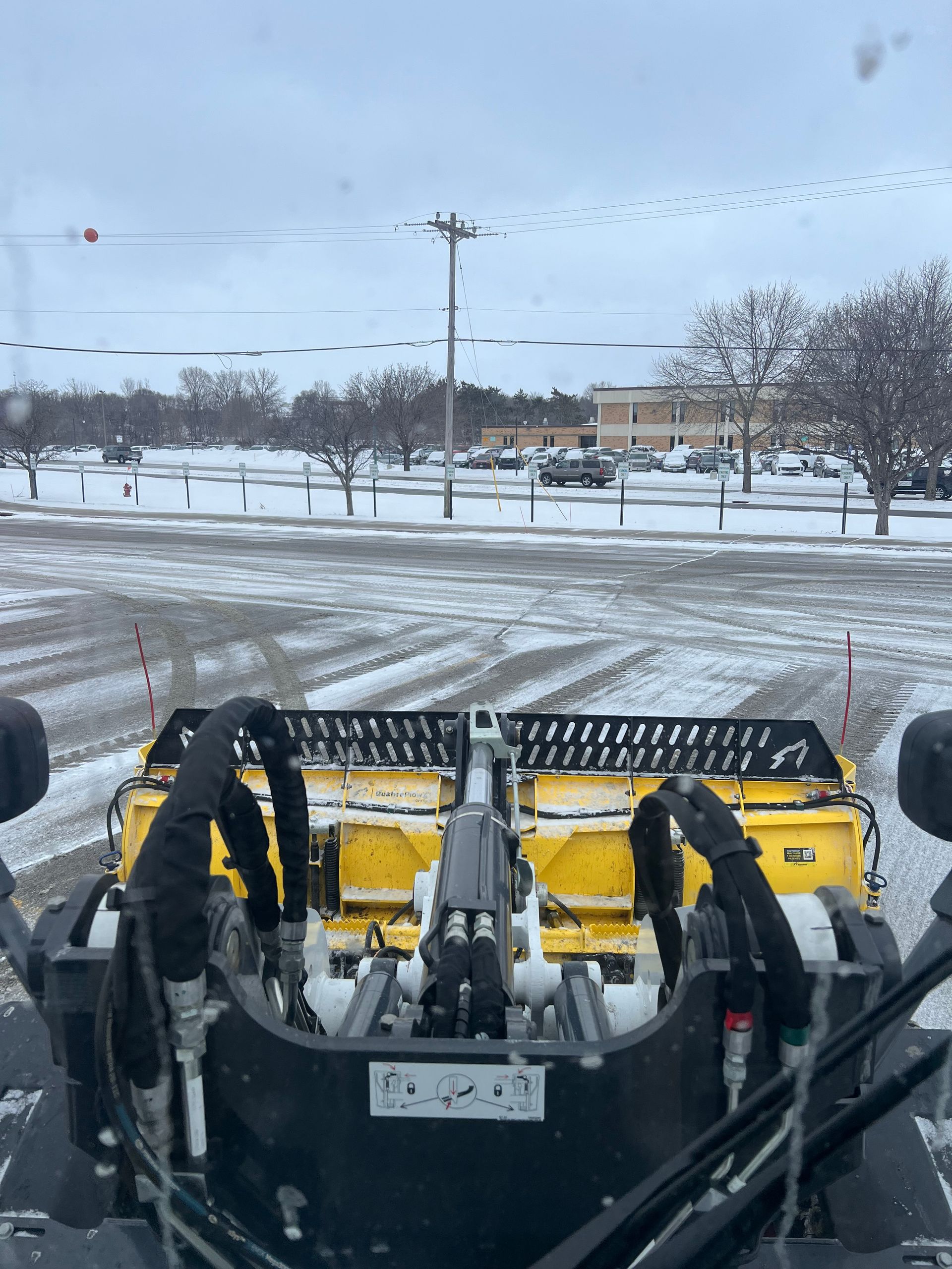 Snowplow blade clearing a snowy parking lot, with winter trees and a road in the background.