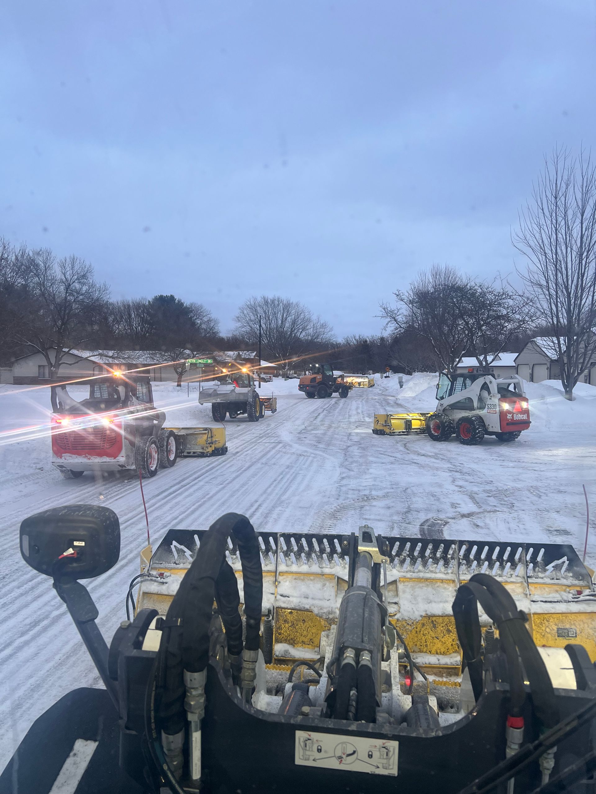 Snowplows clearing a snowy street at dusk, with several vehicles and parked cars in the background
