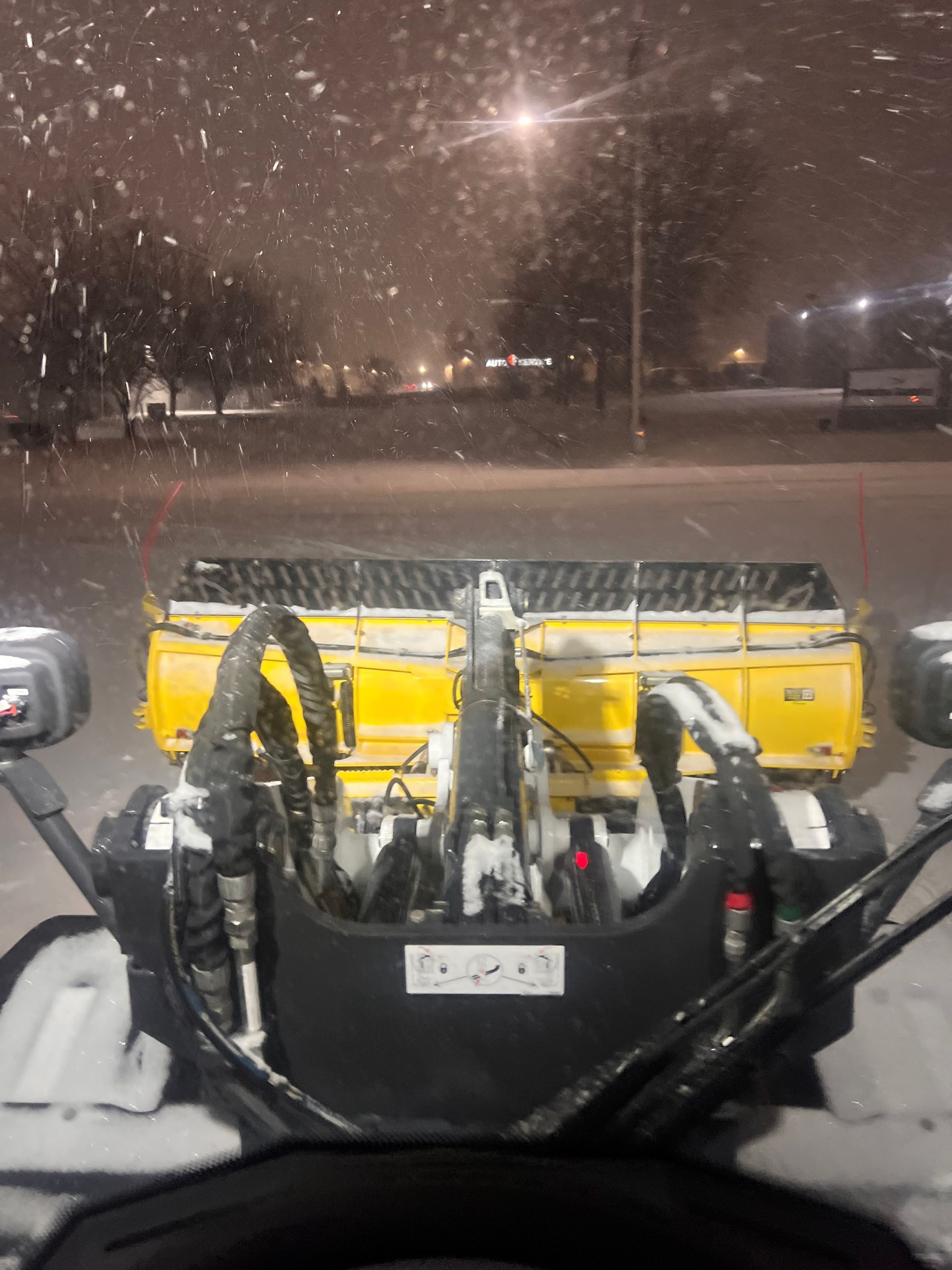 Snowy parking lot with a yellow snowplow loader at night, viewed from inside another vehicle.