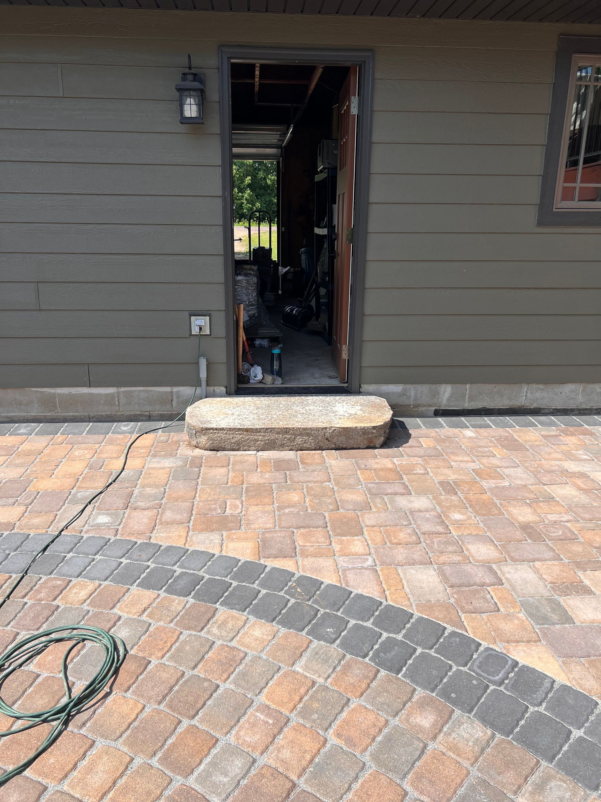 Brick paver patio leading to a dark exterior doorway on a gray house, with a small step at the entrance