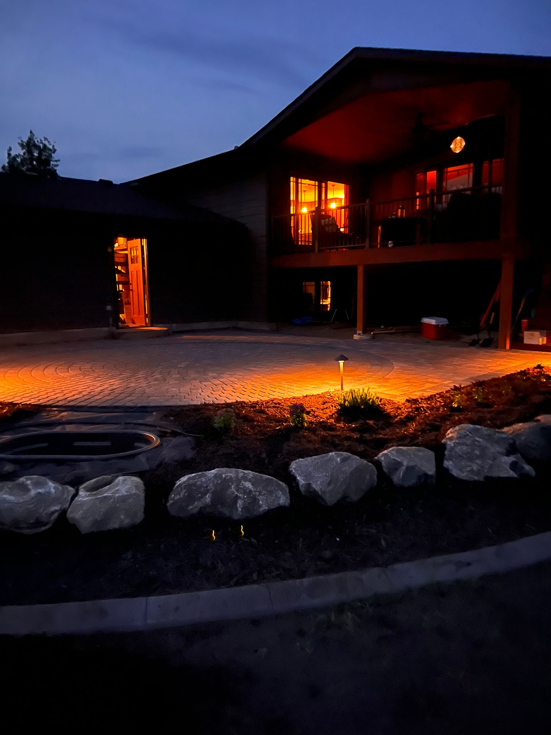 Snowy house at dusk with glowing orange lights and a lit doorway, set beside rocks and a dark driveway.