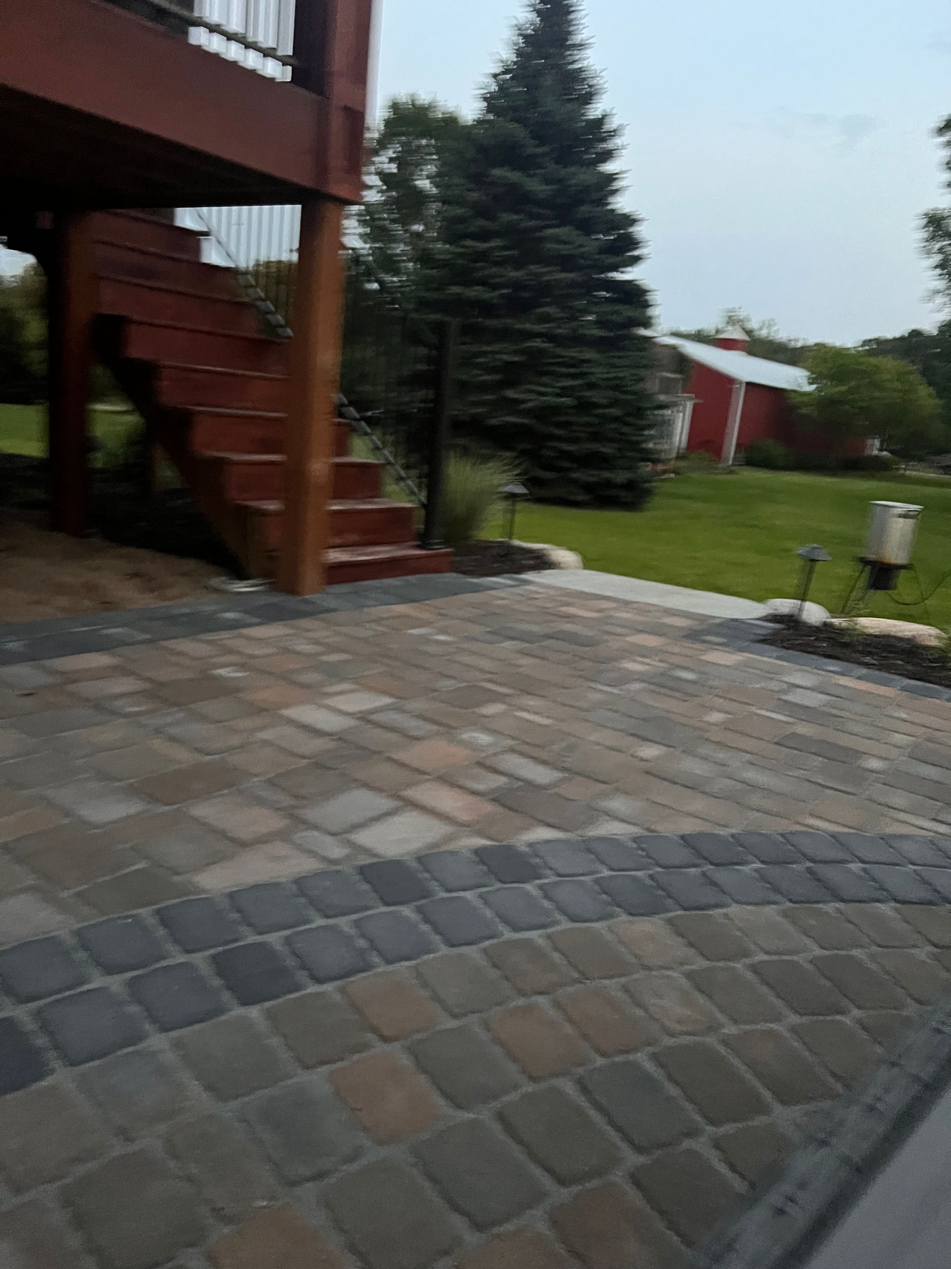 Backyard patio with stone pavers, wooden stairs, evergreen trees, and a grassy lawn after rain