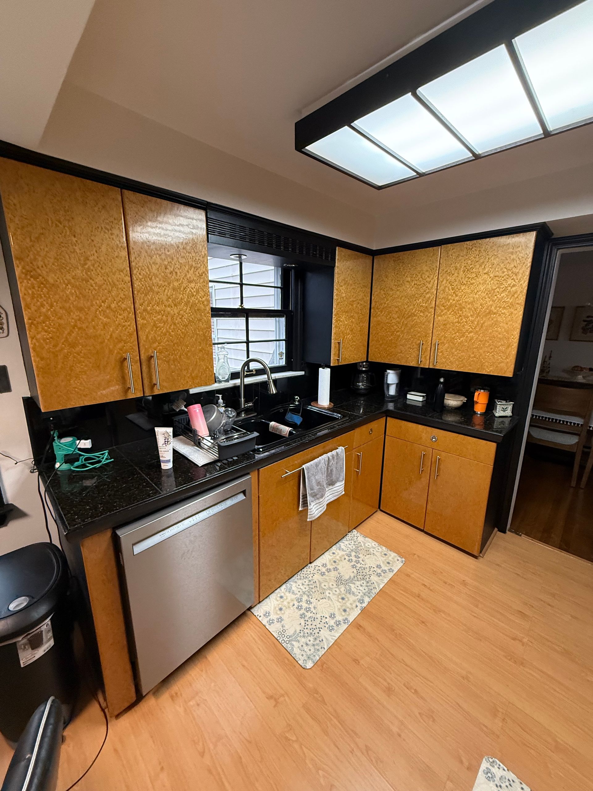 Modern kitchen with black counters, wood cabinets, stainless dishwasher, and skylight ceiling.