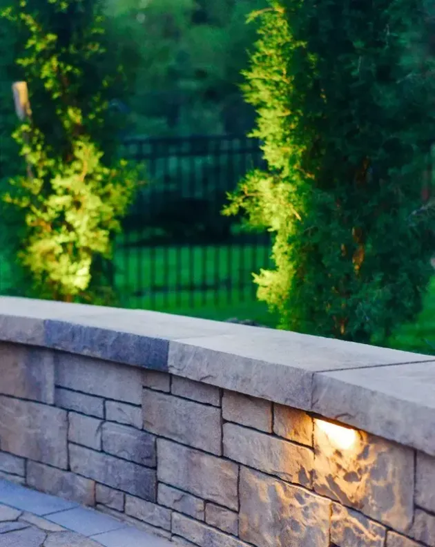 A stone retaining wall with an integrated light glows softly, set against a backdrop of green trees and a black fence.