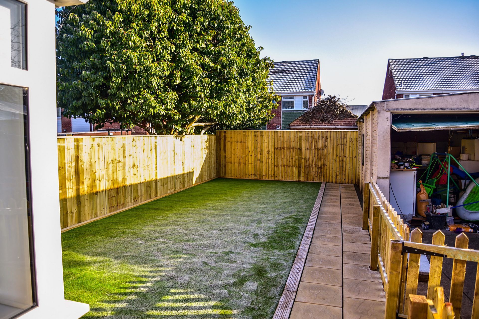 A backyard featuring a fresh wooden fence, a patch of green grass, and a paved stone path leading to a garage structure.