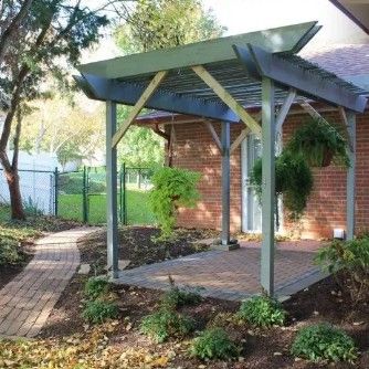 A gray wooden pergola over a brick patio in a backyard with a brick house, hanging plants, and a brick pathway.