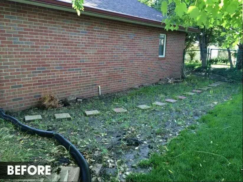 A side yard with a red brick house, a muddy ground area with stepping stones, and a black drainage pipe.