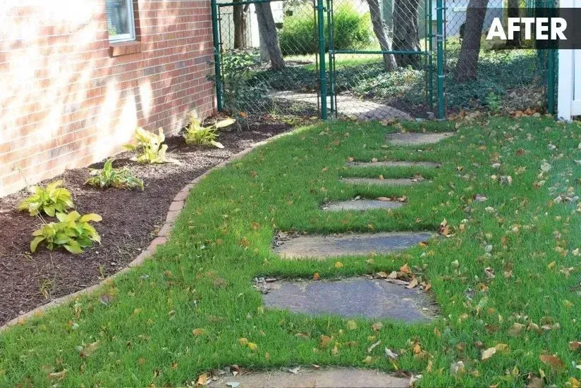 A path of stone stepping stones set into a grassy lawn beside a brick wall and a garden bed with small plants.