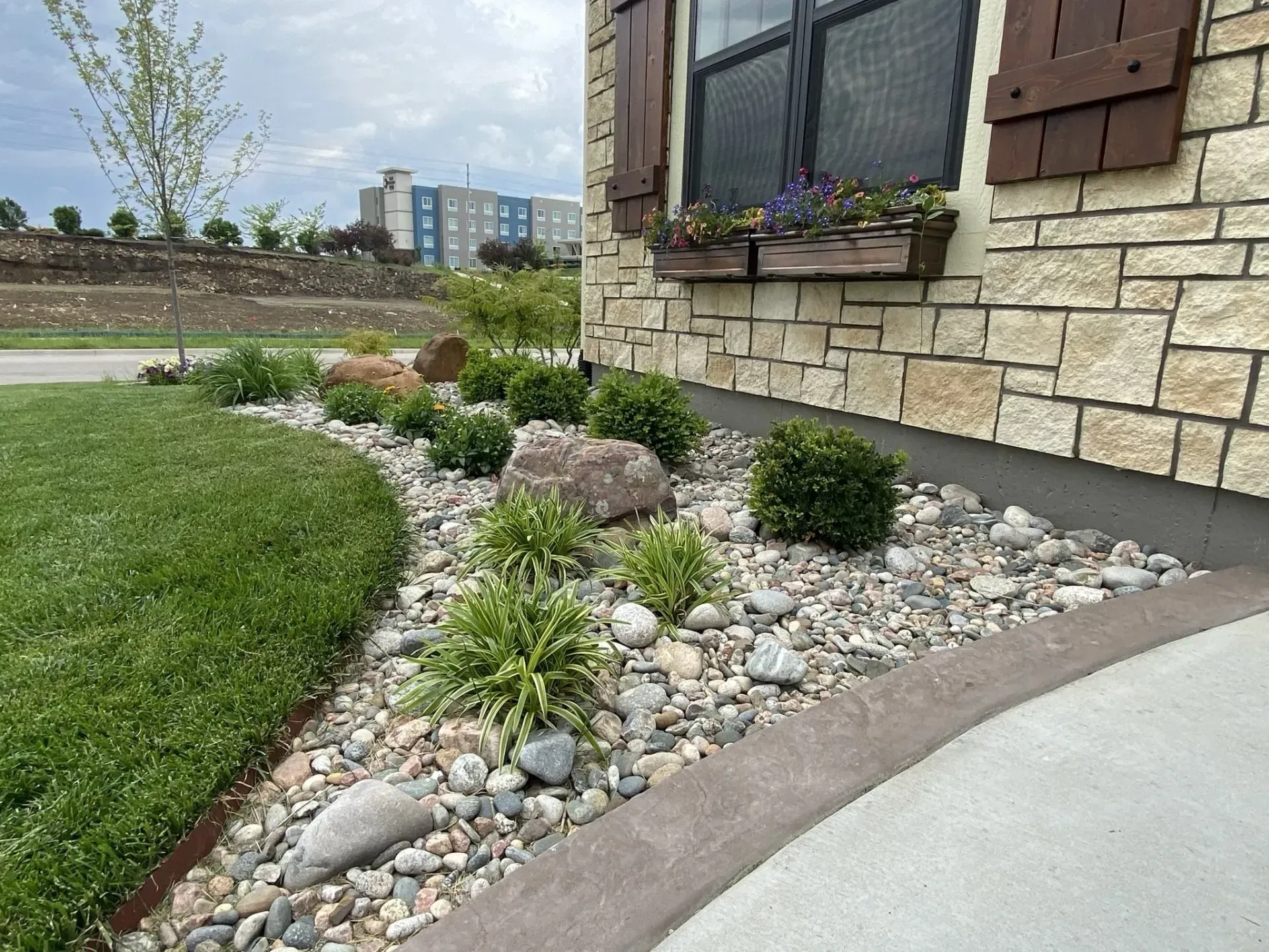 A landscaped garden bed with river stones, small green shrubs, and a boulder next to a stone building wall.